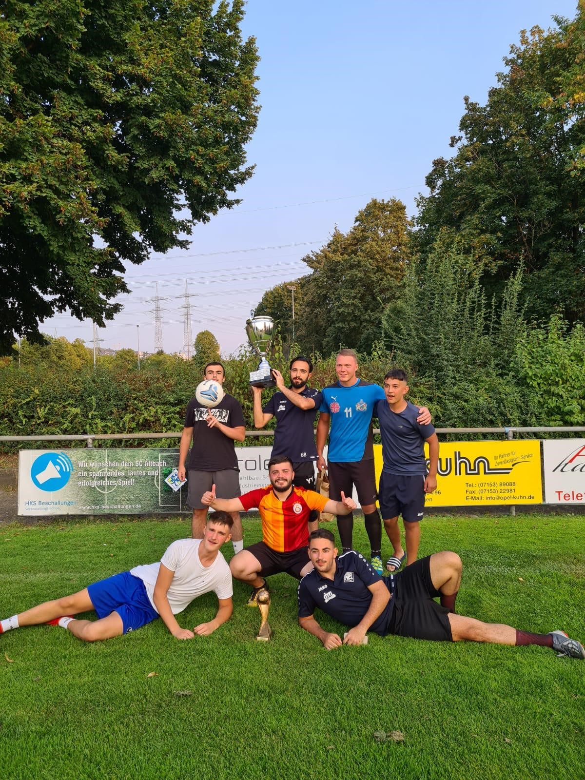 Sieben jubelnde Spieler mit Pokal auf dem Rasen vor Werbebanden des SC Altbach